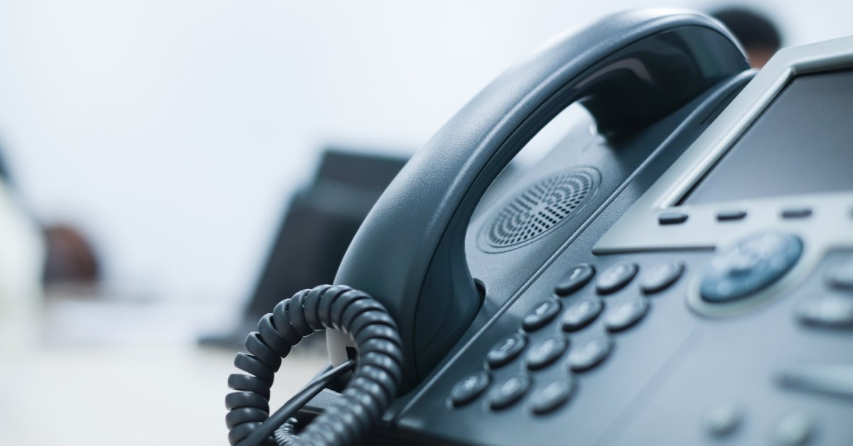 Close-up of a black desk phone with a big handset, dial buttons, a small screen, an exterior speaker, and a cord.