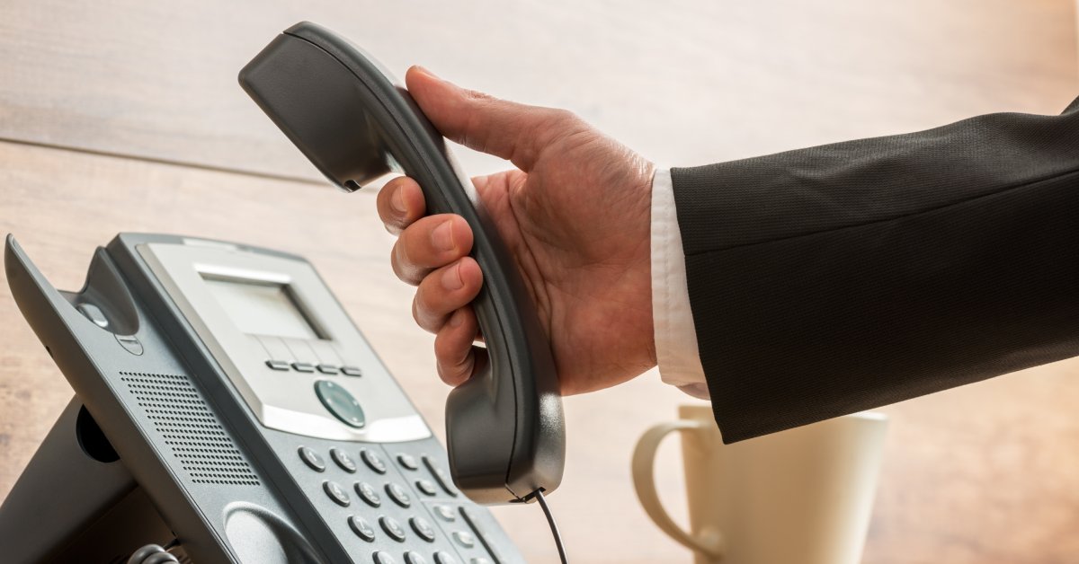 Hand of a man wearing a white shirt and black suit jacket holding the handset with the main system on a desk.