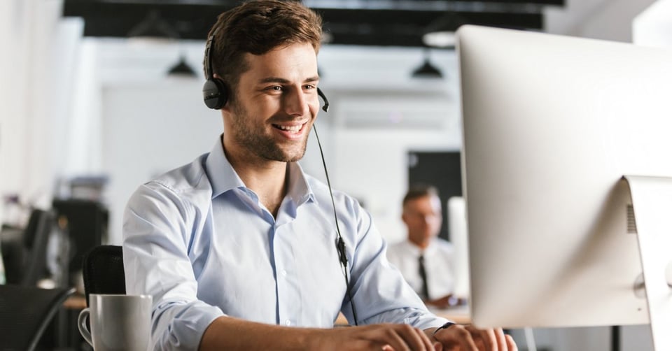 A young professional wearing a button-down shirt and a headset, sitting behind a desk and looking at a monitor.