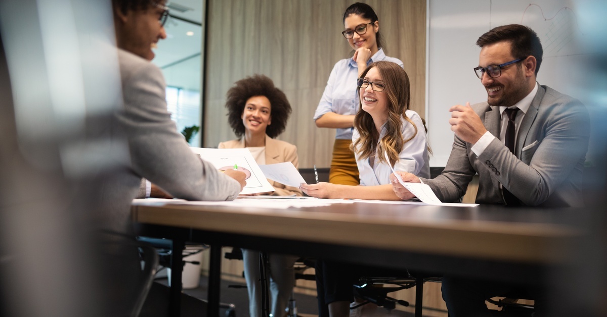 Three women and a man sitting behind a desk, laughing with a man sitting across from them inside a meeting room.