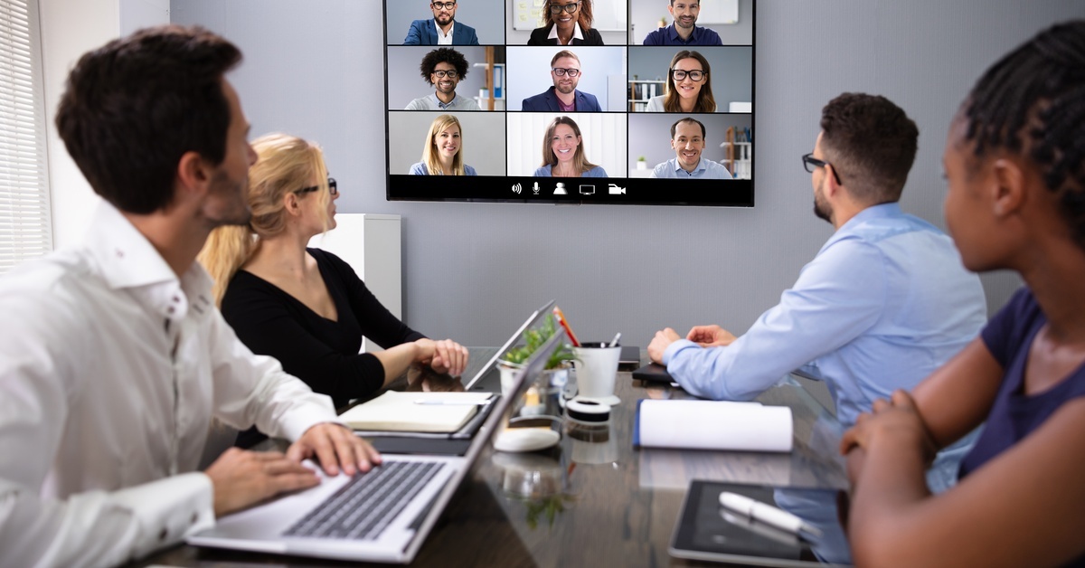 Two women and two men inside a meeting room sitting around a table, looking at a TV screen displaying nine people.