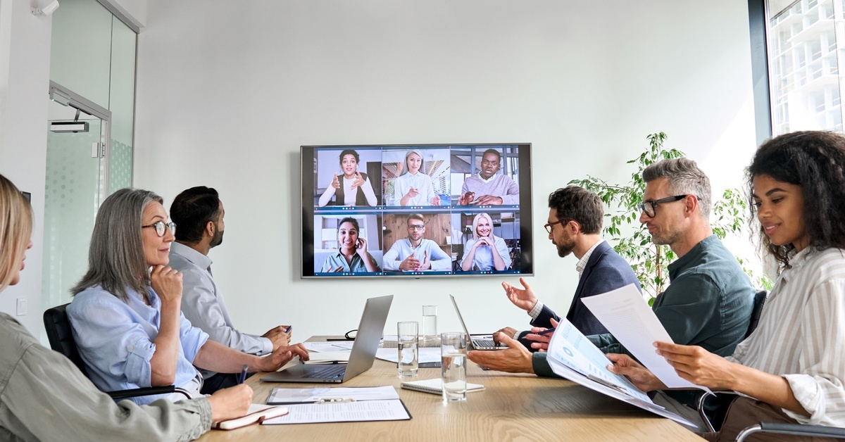 Three women and three men sitting around a desk inside of a meeting room in a video call with six people.