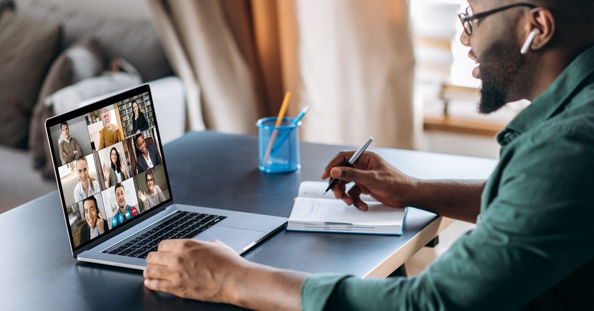 A man sits at a table in a living room. He has a notebook, a pen, and a laptop open to a video call.