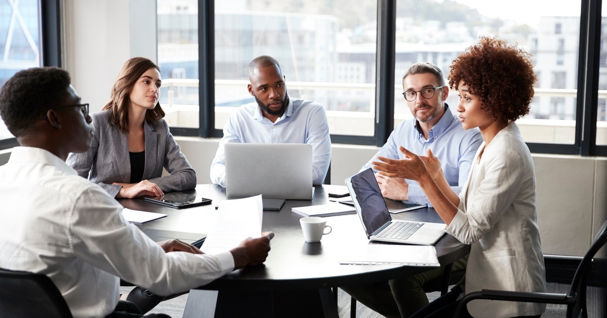 A group of people sit at a table in a small conference room. They have laptops and papers in front of them.