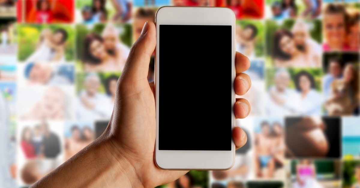 A hand holding a white cell phone that is turned off, set against a vibrant, blurry collage of various people's photos.