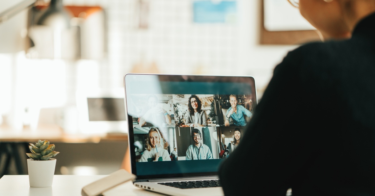 A woman sitting at a neat desk, looking down at her laptop, which displays a video call with several participants.