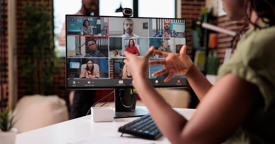A woman sitting at her desk and looking at a monitor showing a video call. She is gesturing with her hands.