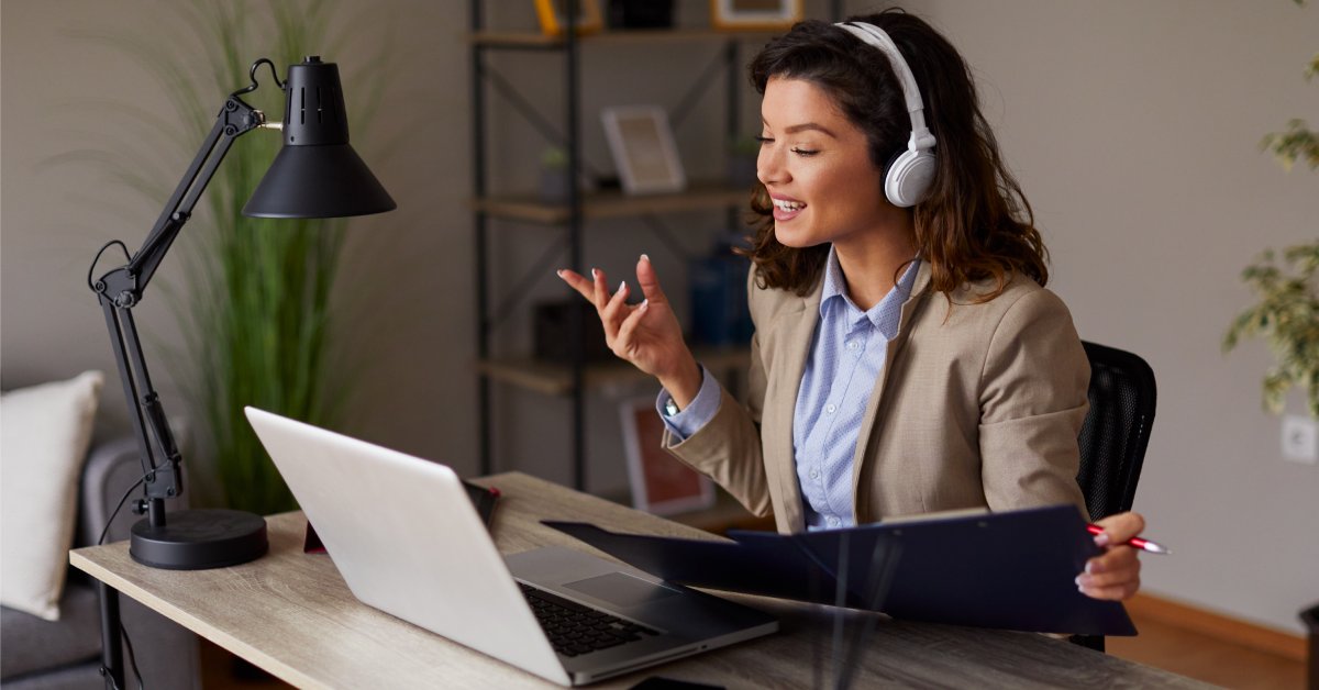 A woman wearing headphones sits at a desk with a laptop. She is holding a folder and looking at the screen.