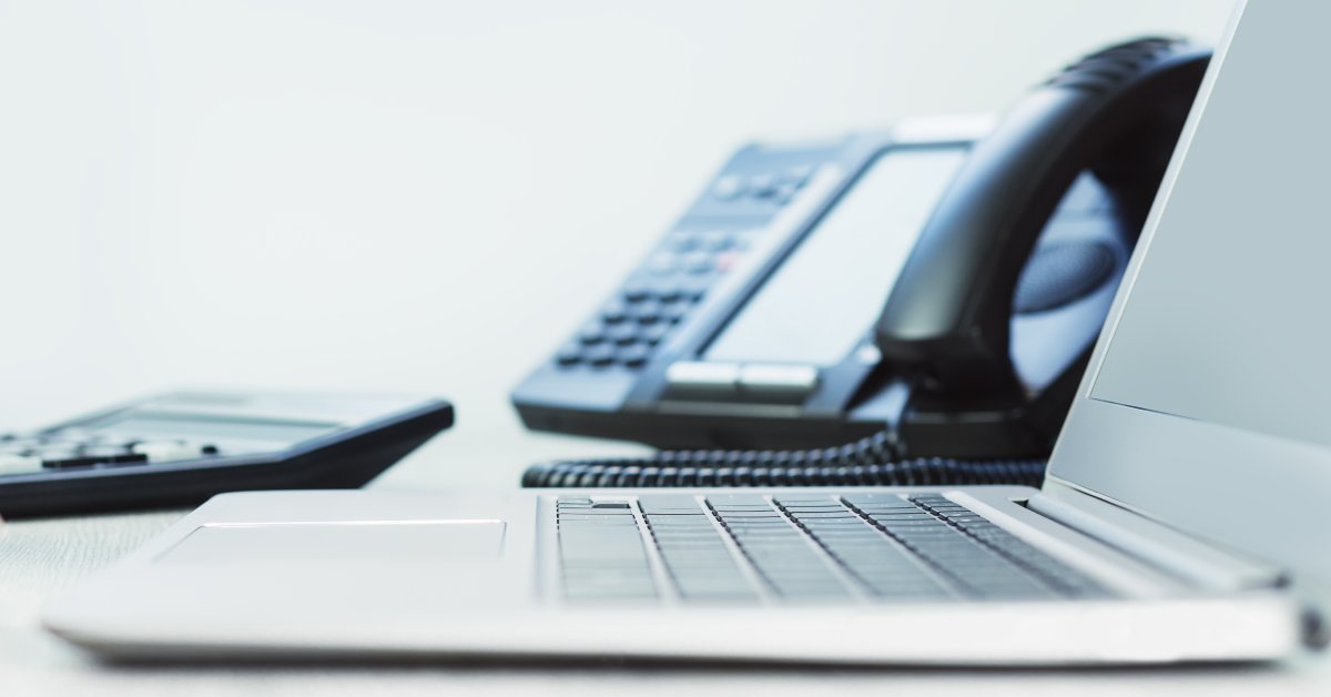 A laptop sitting beside a desk phone and a calculator. The laptop is open but the screen is blank.