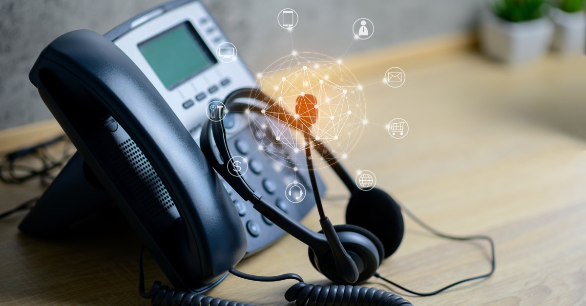 A telephone and headset sitting on a wooden desk. Above the headset mic is a graphic of different communication icons.