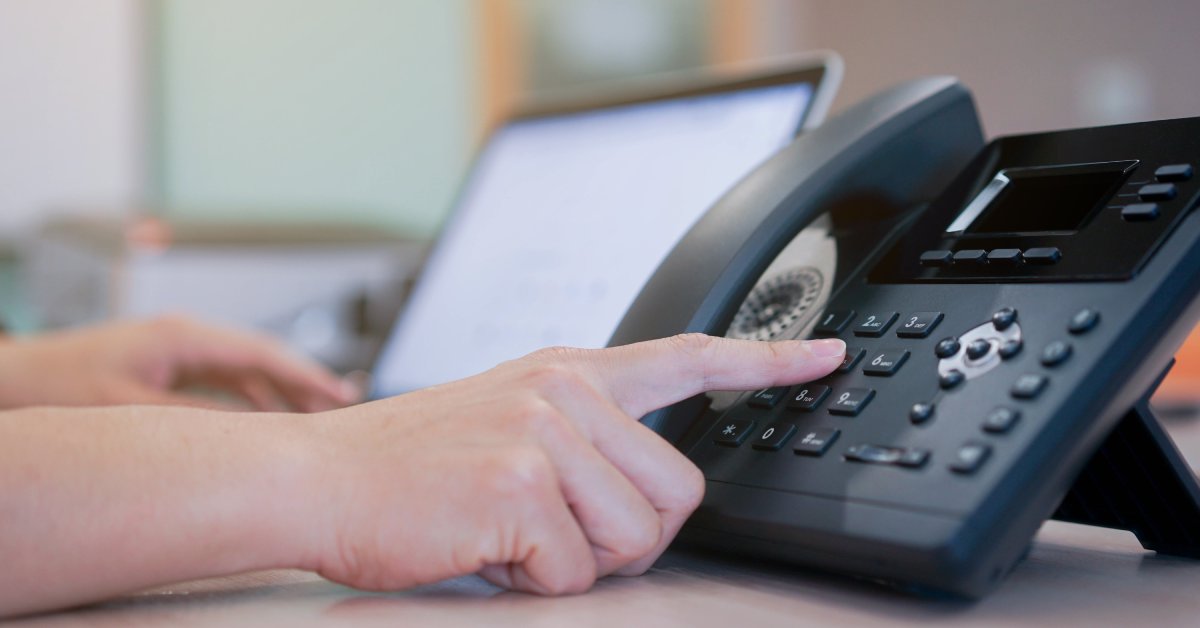 A telephone sitting on a desk next to a laptop. Someone is typing on the laptop while pressing a button on the phone.