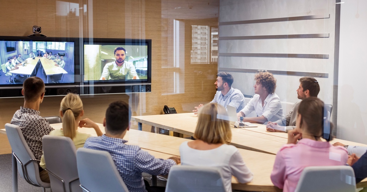 A conference room full of people in business casual wear. They are watching a screen showing a video call.
