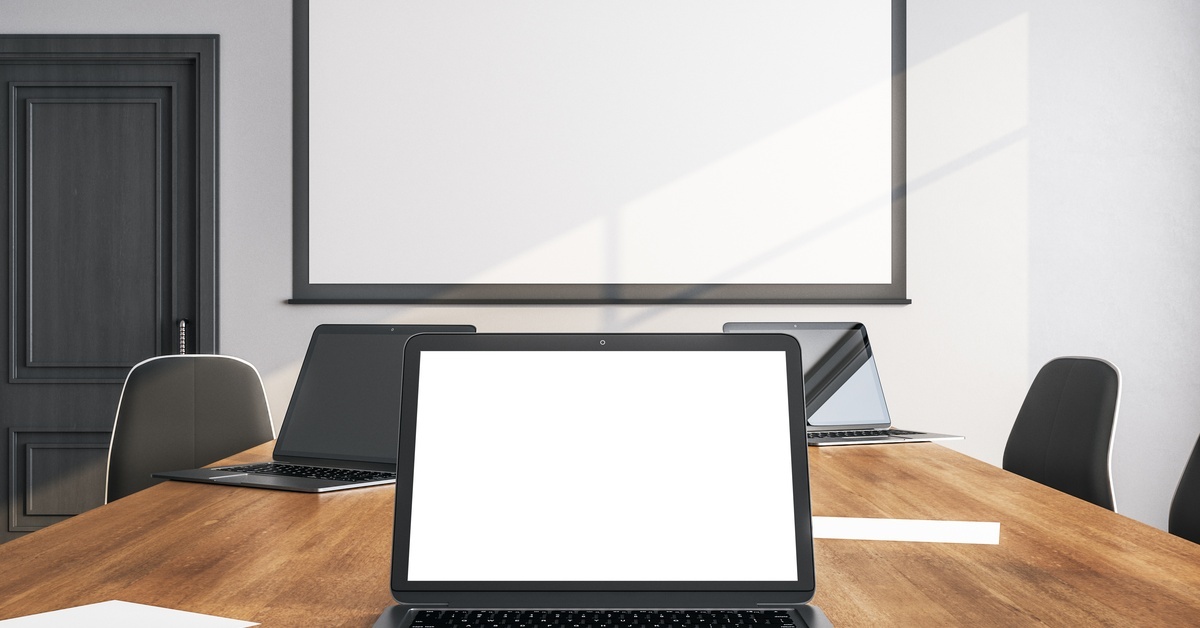 A conference table with blank, open laptops on it. On the wall in front of the table is a white projector screen.