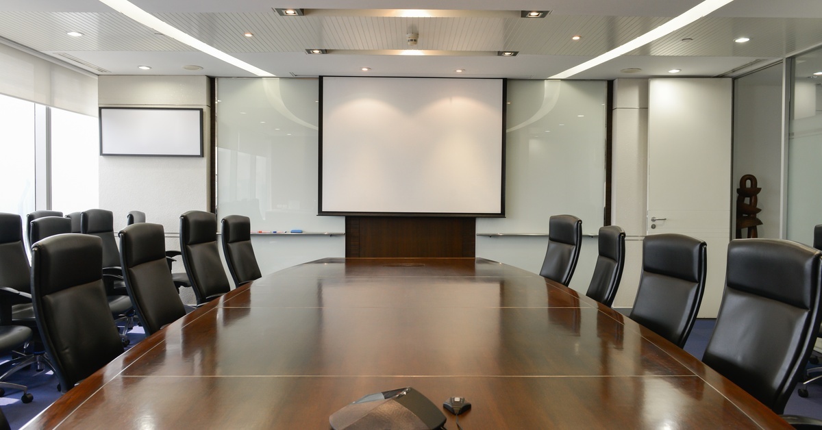 An empty conference room with a long table and office chairs. At the end of the table is a white projector screen.