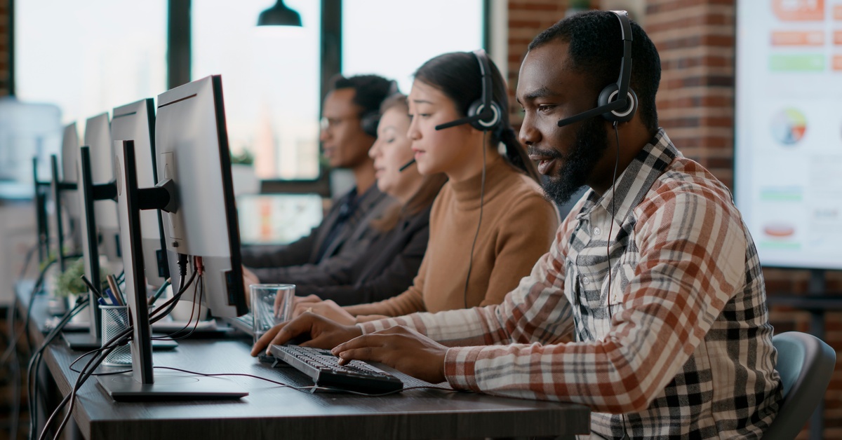 A row of employees in business casual clothing sitting at their monitors with headsets on. They are in a brick-walled room.