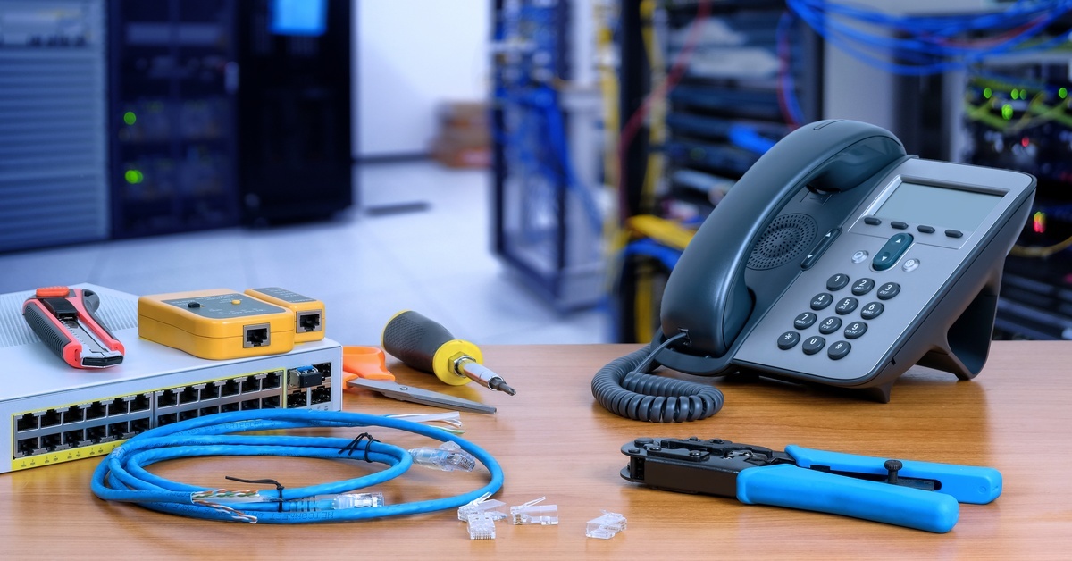 A modern landline sitting on a table with tools, cables, and other various parts. Behind the table is data center equipment.
