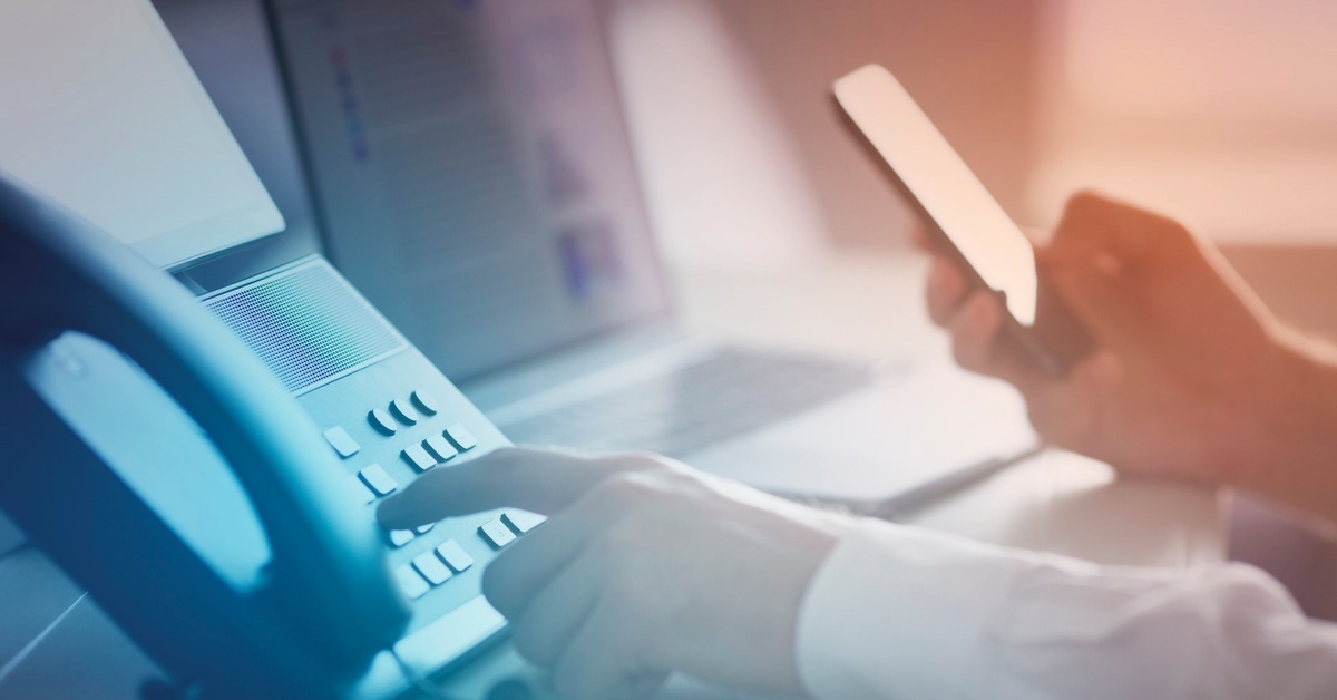 A person holding a mobile phone in one hand and dialing on a landline with the other. There is a laptop on the desk, too.