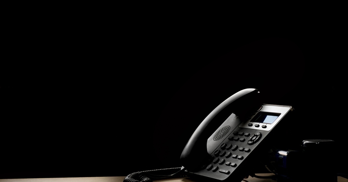 A black modern landline phone sitting on a table with dim, dramatic lighting cast over it creating shadows and highlights.